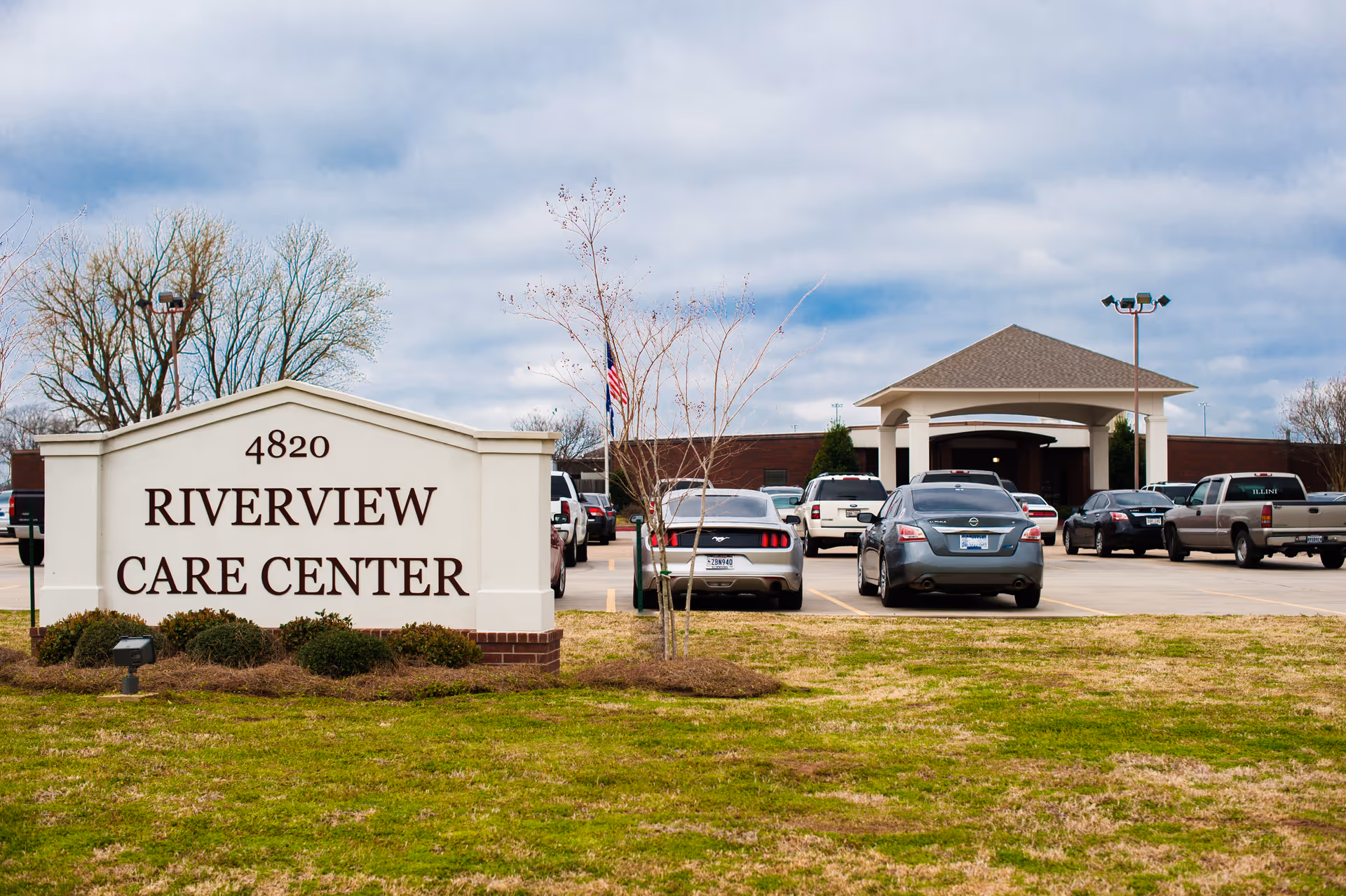 Exterior view of Riverview Care Center showing the entrance with a covered drop-off area, several parked cars, a large sign with the facility name and address, and a grassy area in the foreground under a cloudy sky.