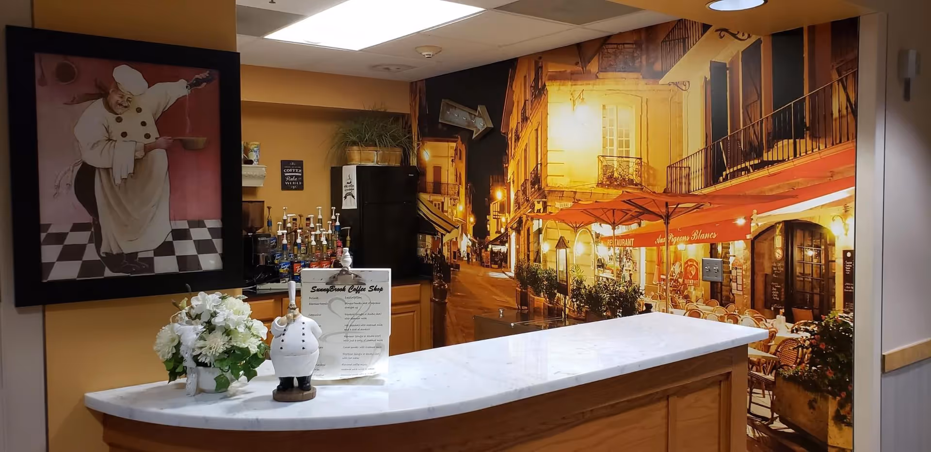 A coffee bar area inside a facility with a marble countertop, a small chef figurine, a flower arrangement, and various coffee syrups. Behind the counter is a large wall mural depicting a warmly lit street scene with outdoor restaurant seating. To the left, there is a framed painting of a chef holding a spoon.