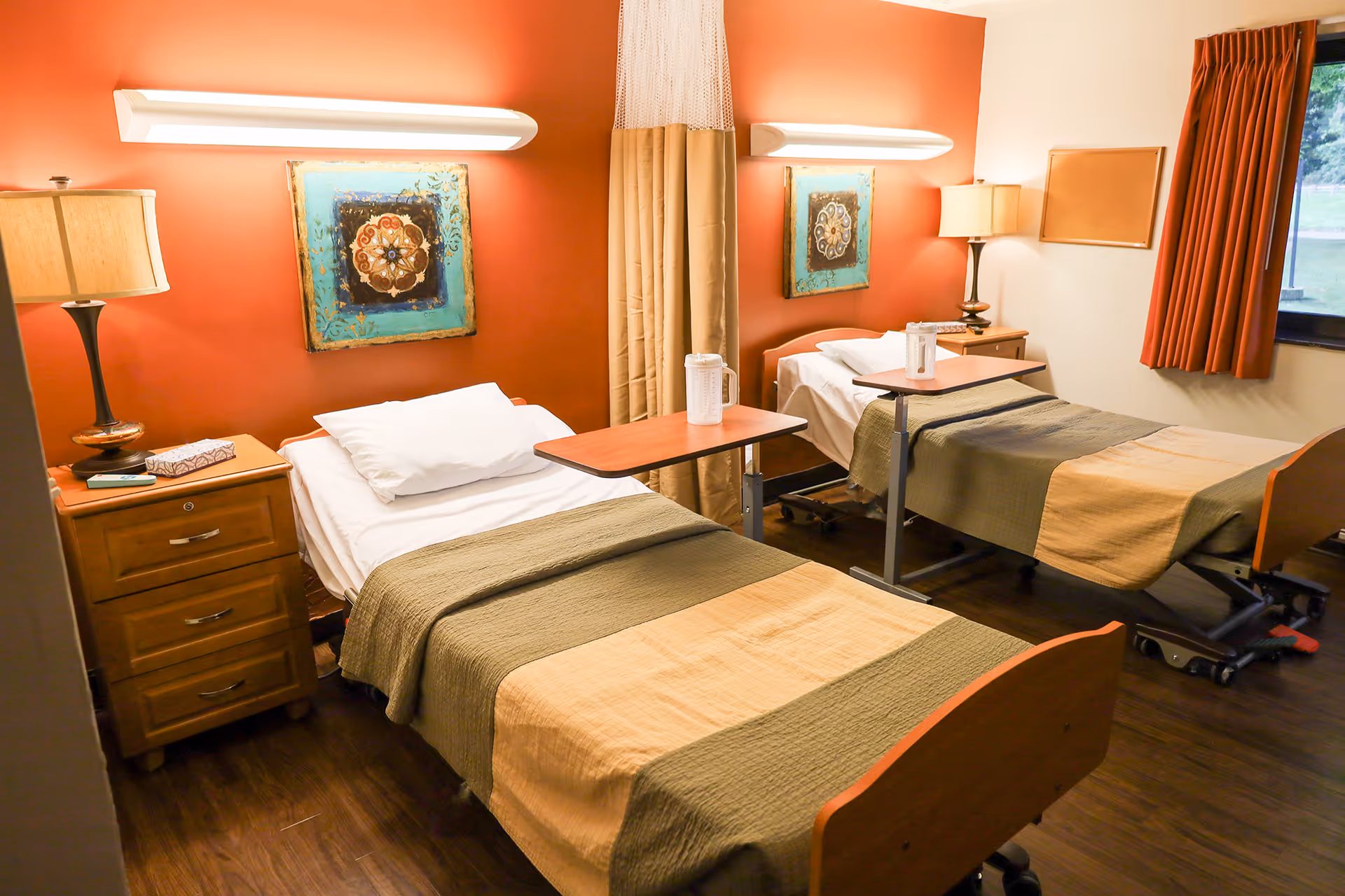 A healthcare facility room with two single beds, each with a beige and green blanket, a white pillow, and an adjustable overbed table with a water pitcher on top. The room has warm orange and beige walls, two bedside tables with lamps, a window with orange curtains, and decorative artwork above each bed.