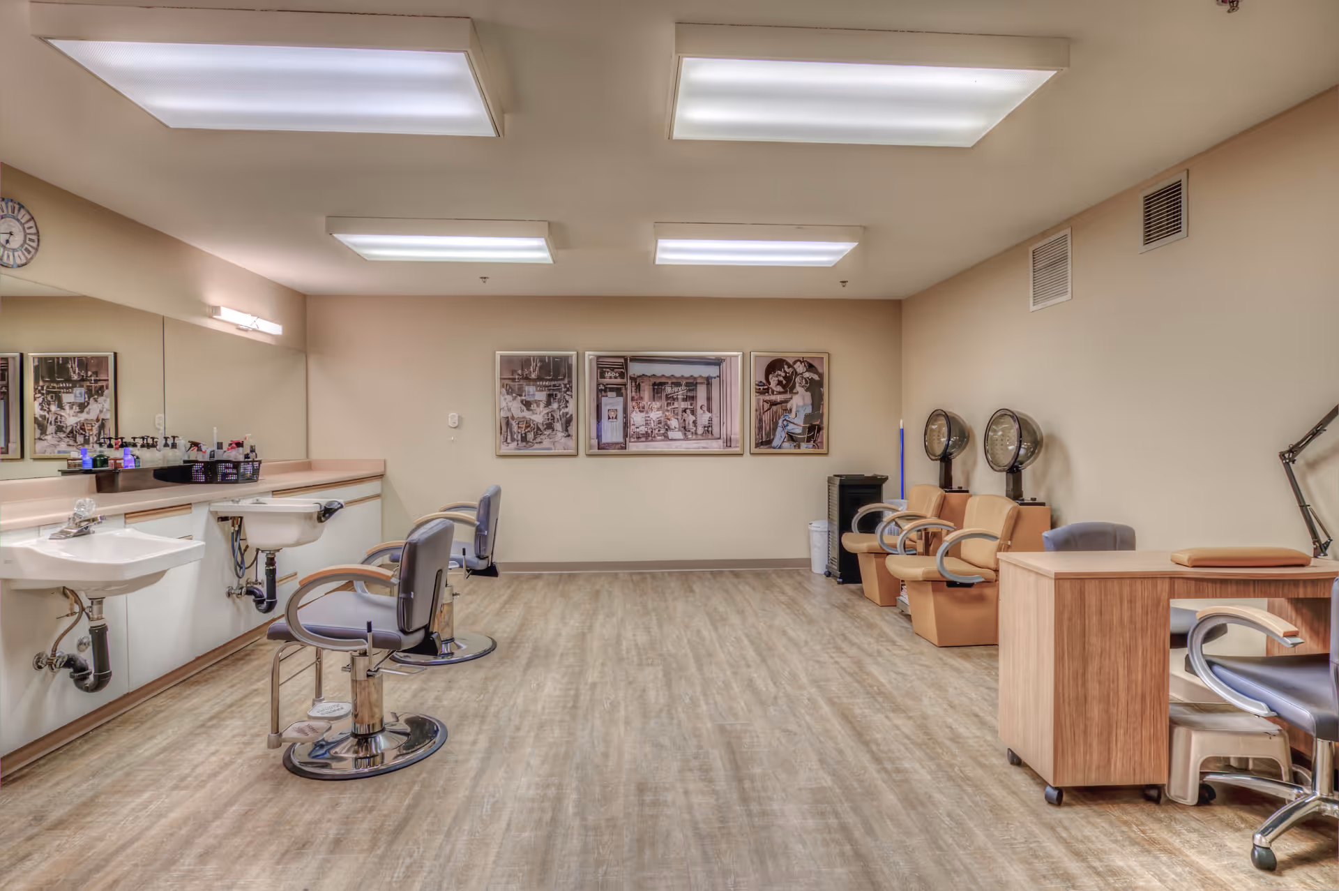 Interior of a salon area in a senior living facility with two salon chairs in front of sinks and mirrors on the left, three beige chairs with hair dryers on the right, a wooden desk with a chair, and framed pictures on the back wall.