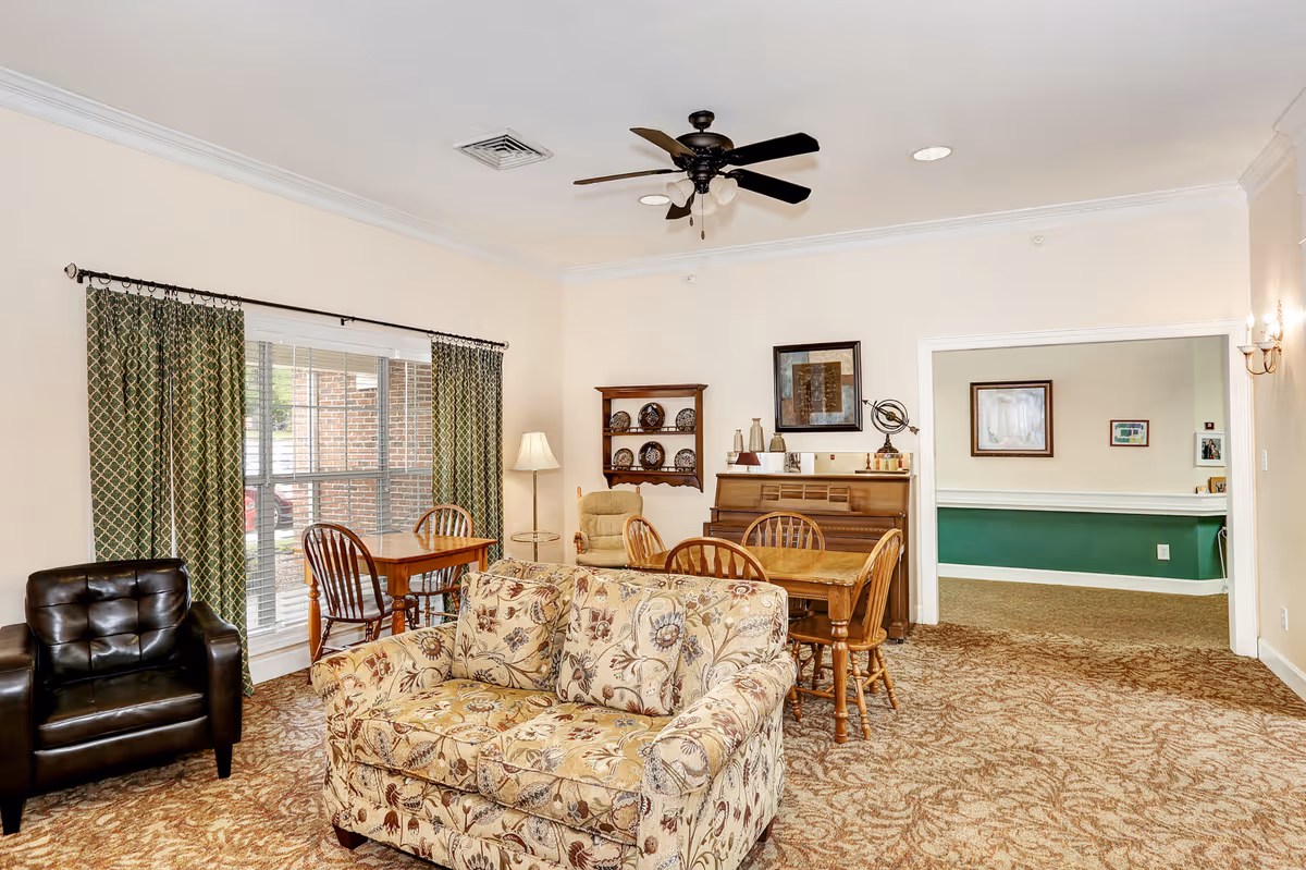 Bright communal living room with a patterned sofa, leather armchair, dining table, piano, and large window with green curtains.
