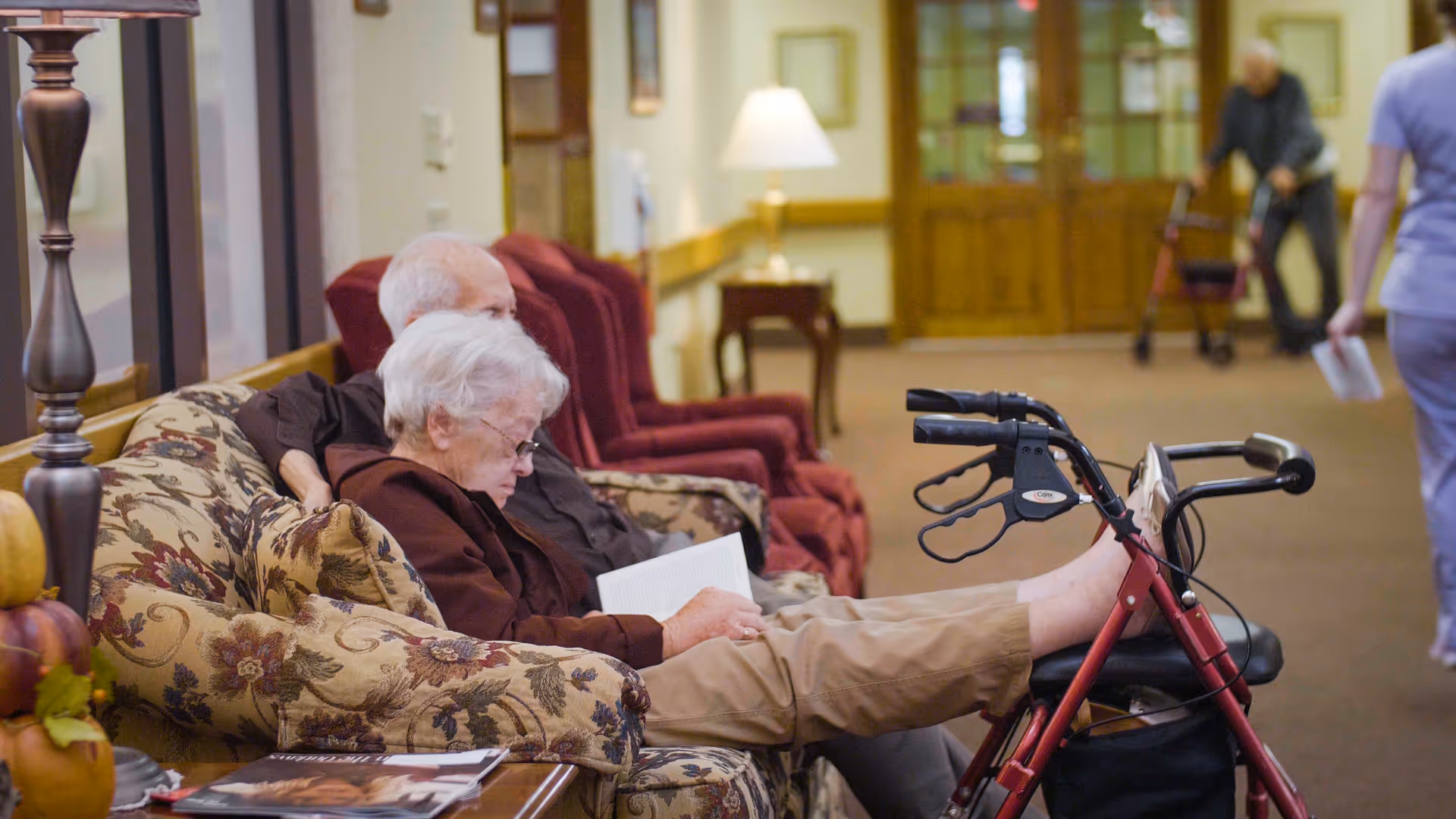 Two elderly residents sit on a floral couch in a facility lounge while one reads a book and a walker is propped nearby.