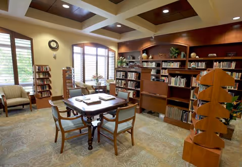 A cozy library room with a wooden table and six chairs in the center. The room has large windows with wooden shutters, a clock on the wall, bookshelves filled with books, and comfortable armchairs near the windows. The ceiling has recessed lighting and decorative wooden beams.