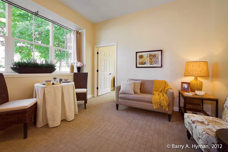 A cozy living room area with a small round table covered with a white tablecloth, two wicker chairs, a beige loveseat with pillows and a yellow throw blanket, a side table with a lamp and framed photo, and a floral armchair. Large windows show green trees outside, and a door is open to another room.