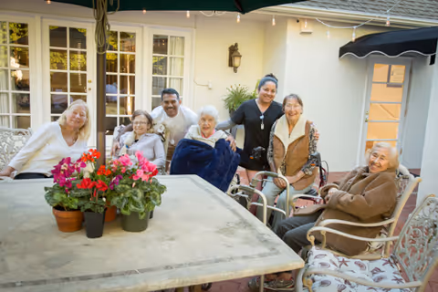 A group of six elderly individuals and one caregiver sitting and standing around an outdoor patio table with potted flowers, smiling and enjoying each other's company in a courtyard area with string lights and a building with windows in the background.