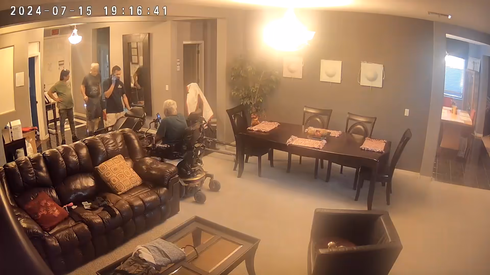 A living room and dining area in a senior assisted living home. The living room has a dark brown leather couch with pillows and a glass coffee table. The dining area has a wooden table with four chairs and placemats. There are four people standing near a doorway and one person sitting in a motorized wheelchair. The walls are painted gray with three decorative wall hangings and a potted plant. A bright ceiling light illuminates the room.