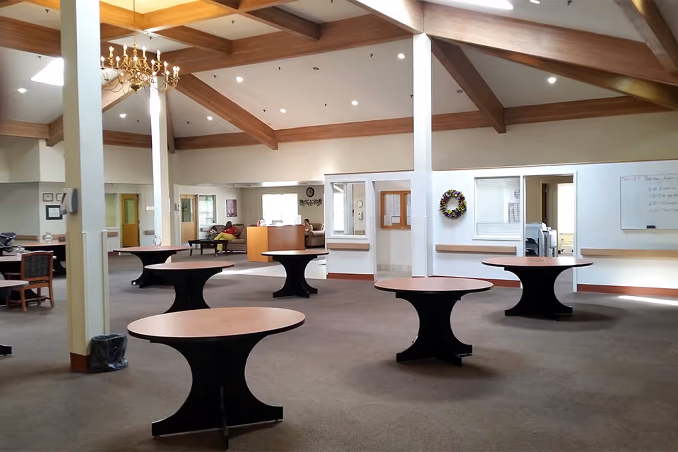 A spacious interior room with a high ceiling featuring wooden beams and a chandelier. The room contains several round tables with black bases and wooden tops spaced evenly across a carpeted floor. In the background, there is a reception desk with a person sitting behind it, and seating areas with couches and chairs. The walls are light-colored with some windows and a whiteboard visible.