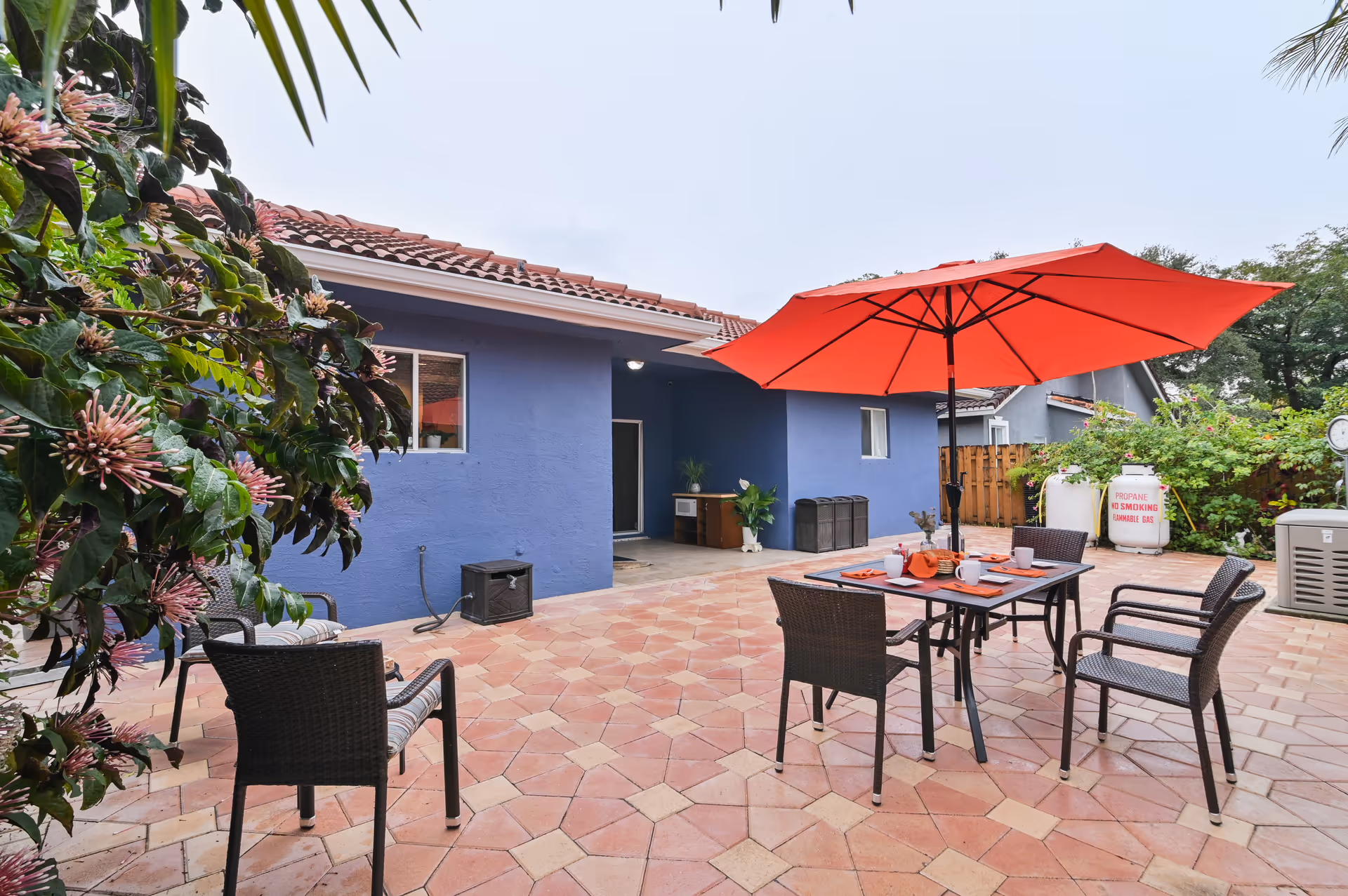 Outdoor patio area with a tiled floor, featuring a table with four chairs and a large red umbrella. The patio is adjacent to a blue building with a tiled roof. There are plants and greenery around the patio, including a flowering bush on the left side. A propane tank with a 'No Smoking' sign is visible in the background.