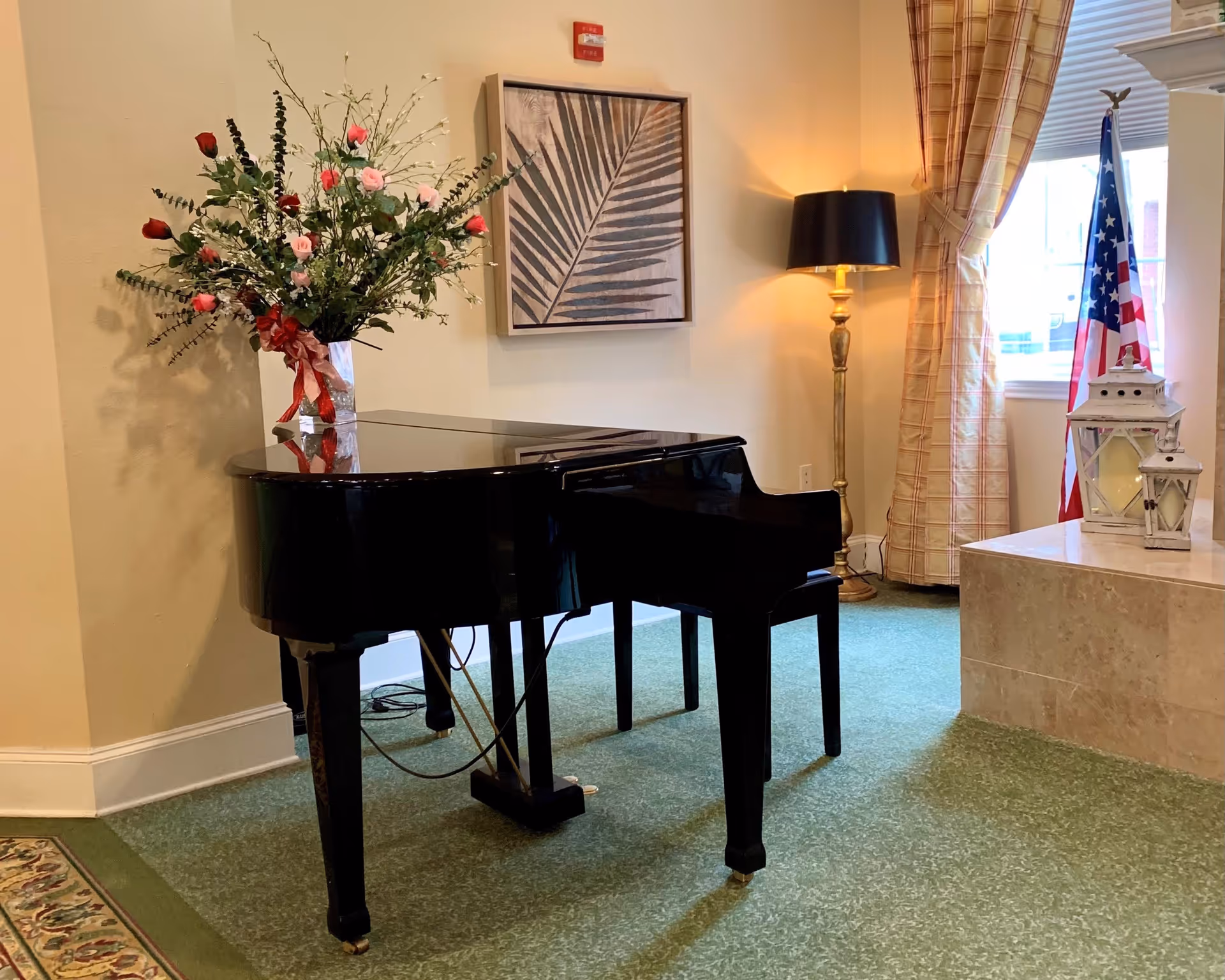 A black grand piano with a vase of flowers on top is placed in a room with green carpet. There is a framed artwork of a leaf on the wall behind the piano, a tall floor lamp with a black shade, plaid curtains, an American flag, and two white lanterns on a marble ledge near a window.