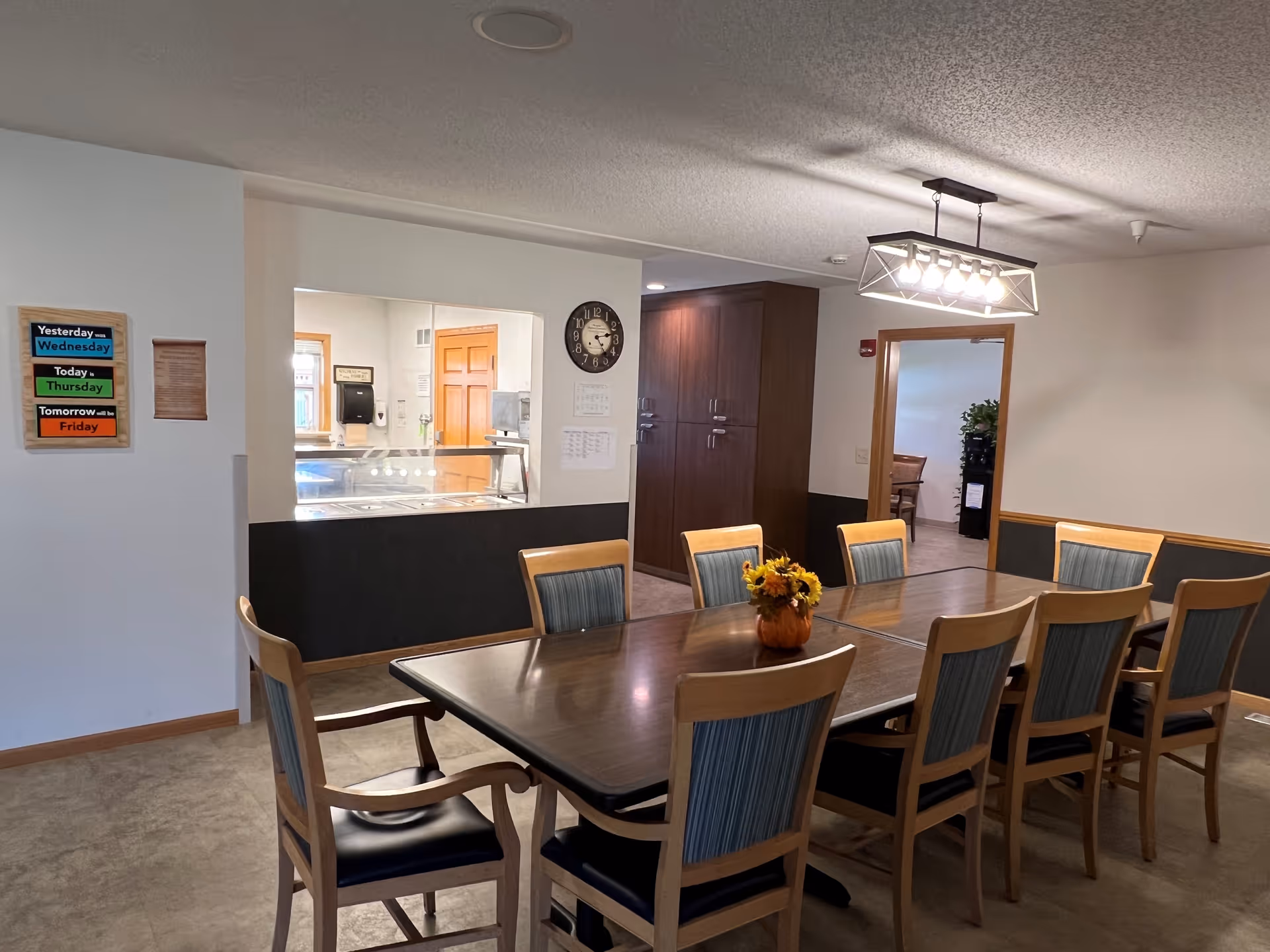 A dining room with a long wooden table surrounded by eight chairs with wooden frames and blue cushioned backs and seats. A small pumpkin-shaped vase with yellow flowers is placed in the center of the table. On the wall to the left, there is a colorful sign showing the days of the week and a clock above a pass-through window to a kitchen area. The room has beige walls with a dark wainscot and a modern rectangular ceiling light fixture. A doorway leads to another room with more chairs and a plant visible.
