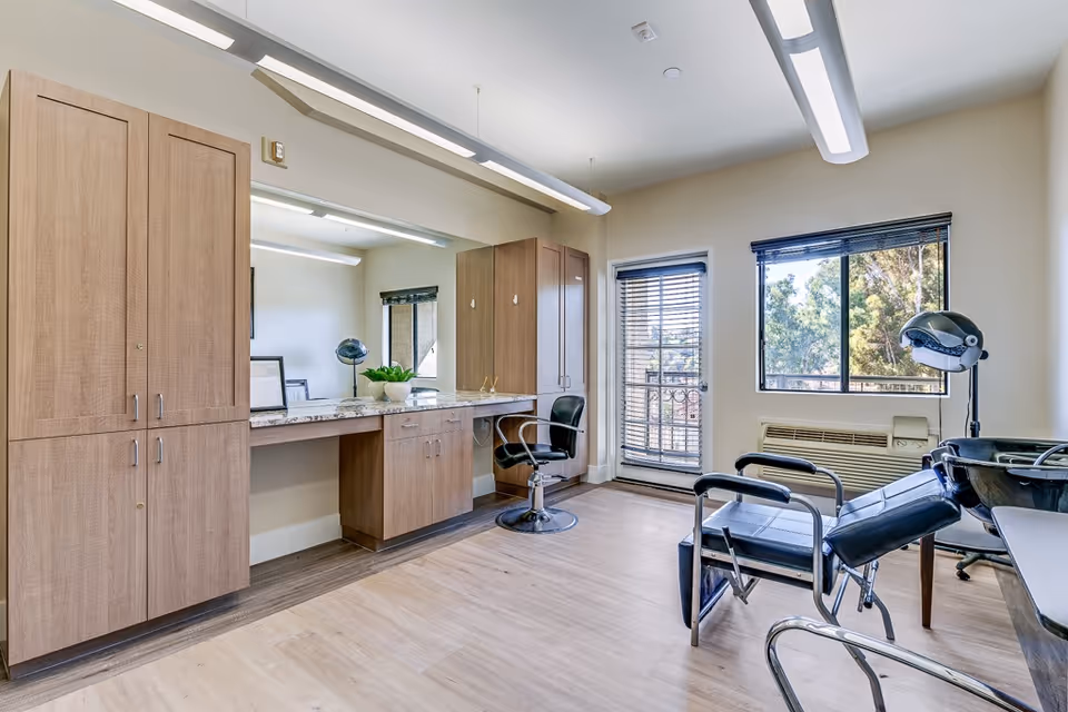 A bright and clean salon room with wooden cabinets, a large mirror, a styling chair, a hair washing station, and a window with blinds letting in natural light.