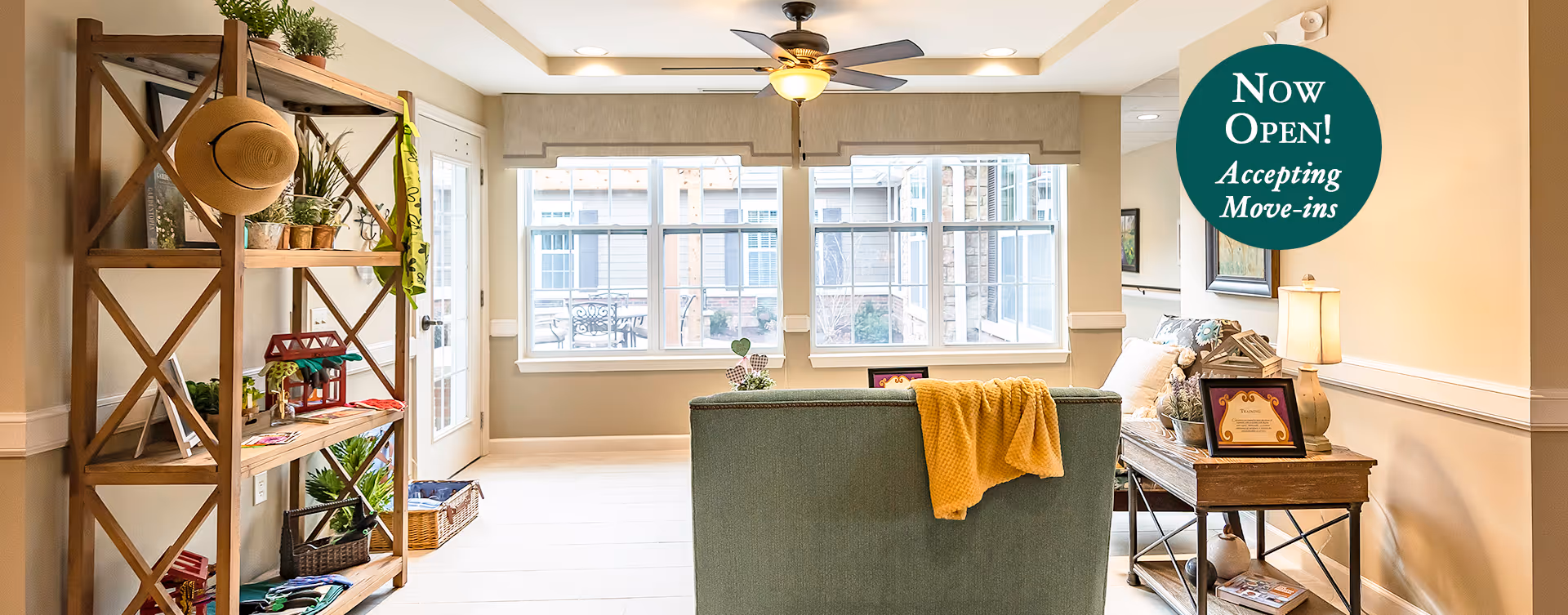 A bright and cozy living room area with a green upholstered chair draped with a yellow blanket, a wooden shelving unit with plants and decorative items on the left, and a wooden side table with a lamp and framed pictures on the right. Large windows let in natural light, and a ceiling fan with a light fixture is mounted on the ceiling. A door leads to an outdoor patio area with a table and chairs visible through the windows.