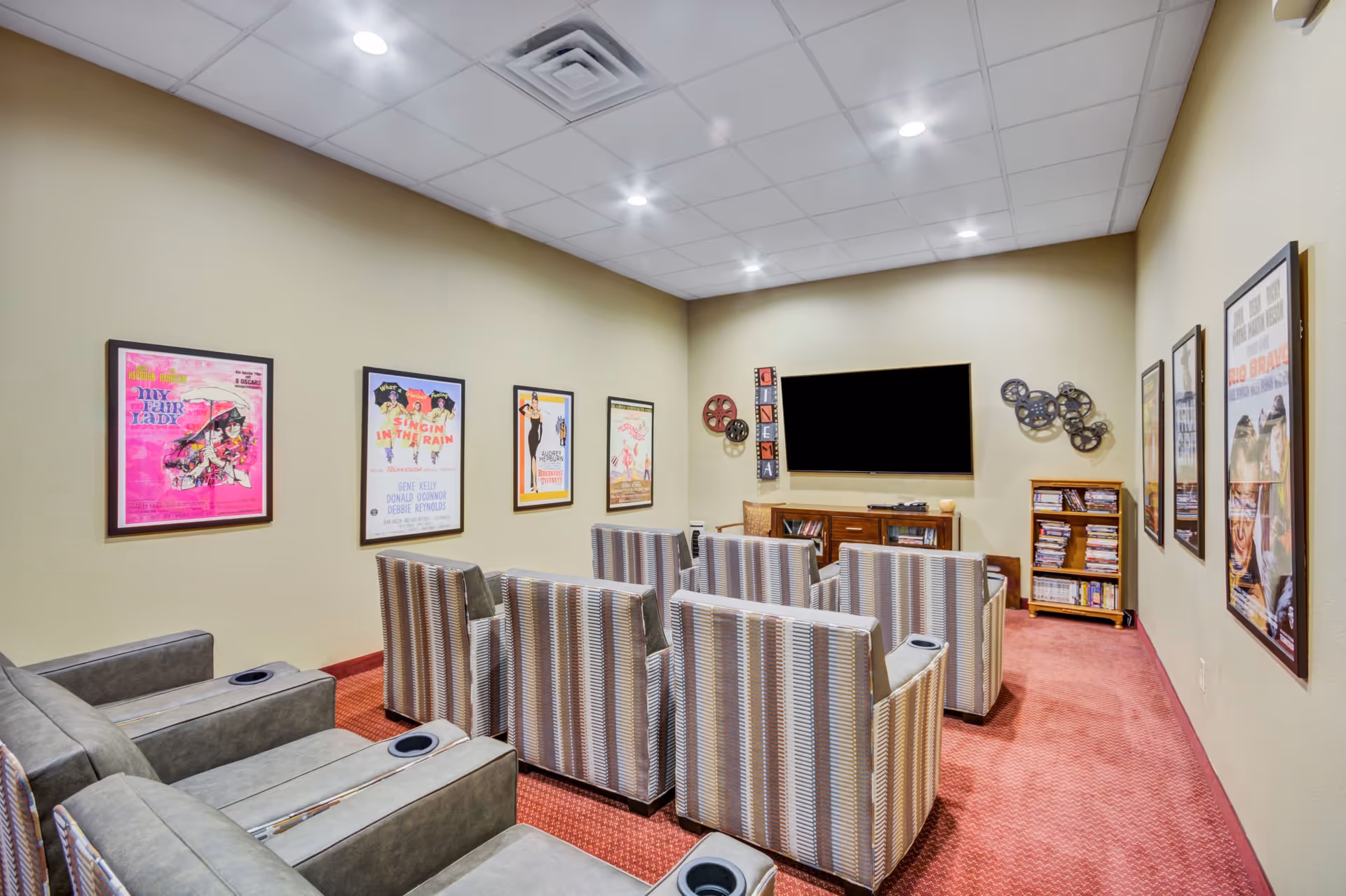 A cozy media room with multiple comfortable armchairs arranged in rows facing a large flat-screen TV mounted on the wall. The walls are decorated with framed vintage movie posters and film reel decorations. There is a small wooden cabinet and a bookshelf filled with DVDs beneath the TV. The room has a carpeted floor and a drop ceiling with recessed lighting.