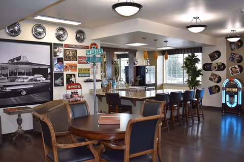 A retro-themed common area with a round wooden table and chairs in the foreground, a bar counter with high stools, vintage signs and hubcaps on the walls, a large black and white photo of a classic car, and a jukebox near the window with plants and natural light coming through.