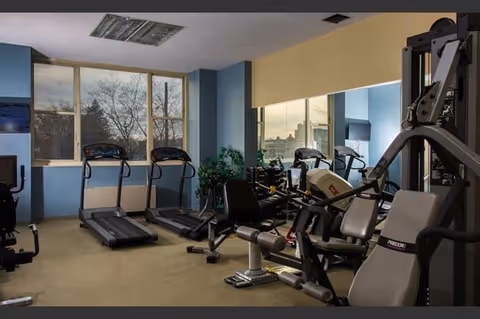 Indoor fitness room with exercise equipment including treadmills, stationary bikes, and weight machines. Large windows provide natural light and a view of trees outside. The walls are painted blue and yellow, and there is a large mirror on one wall.