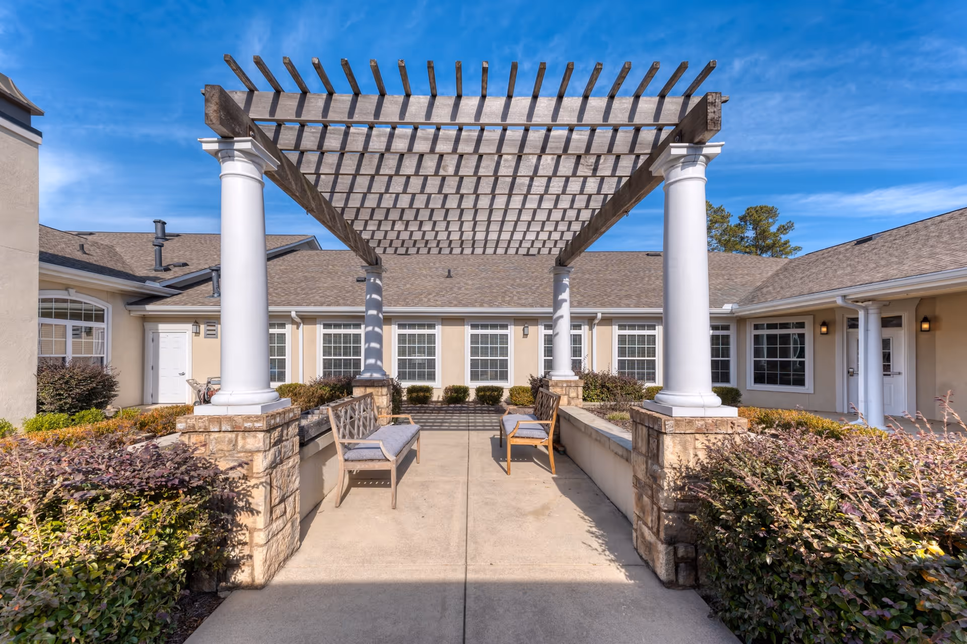 Outdoor courtyard area at Addington Place Of Alpharetta featuring a pergola with white columns, benches with cushions, surrounded by bushes and the exterior walls and windows of the building under a clear blue sky.