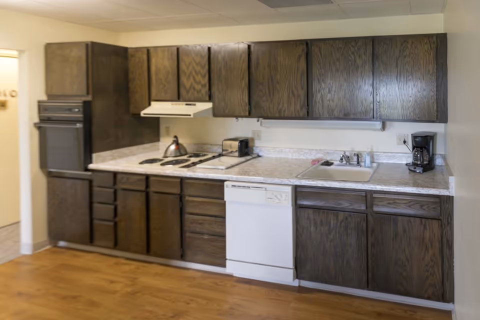 A kitchen area with dark wooden cabinets above and below a white countertop. The countertop includes a sink with a faucet, a coffee maker, a toaster, a stovetop with a kettle, and a dishwasher. The floor is wooden, and the walls are light-colored.