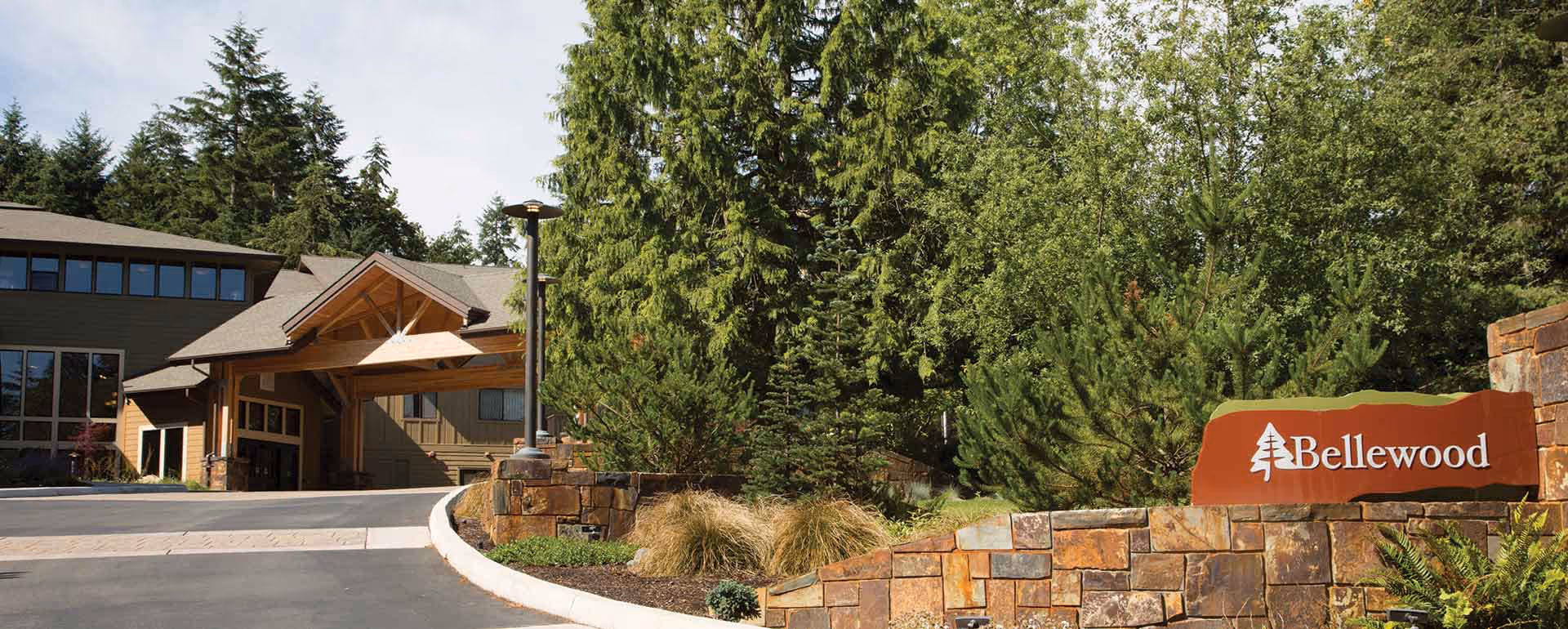 Exterior view of Bellewood - A LifeMinded Residence, showing the entrance to the building surrounded by trees and landscaping with a stone wall and a sign that reads 'Bellewood'.
