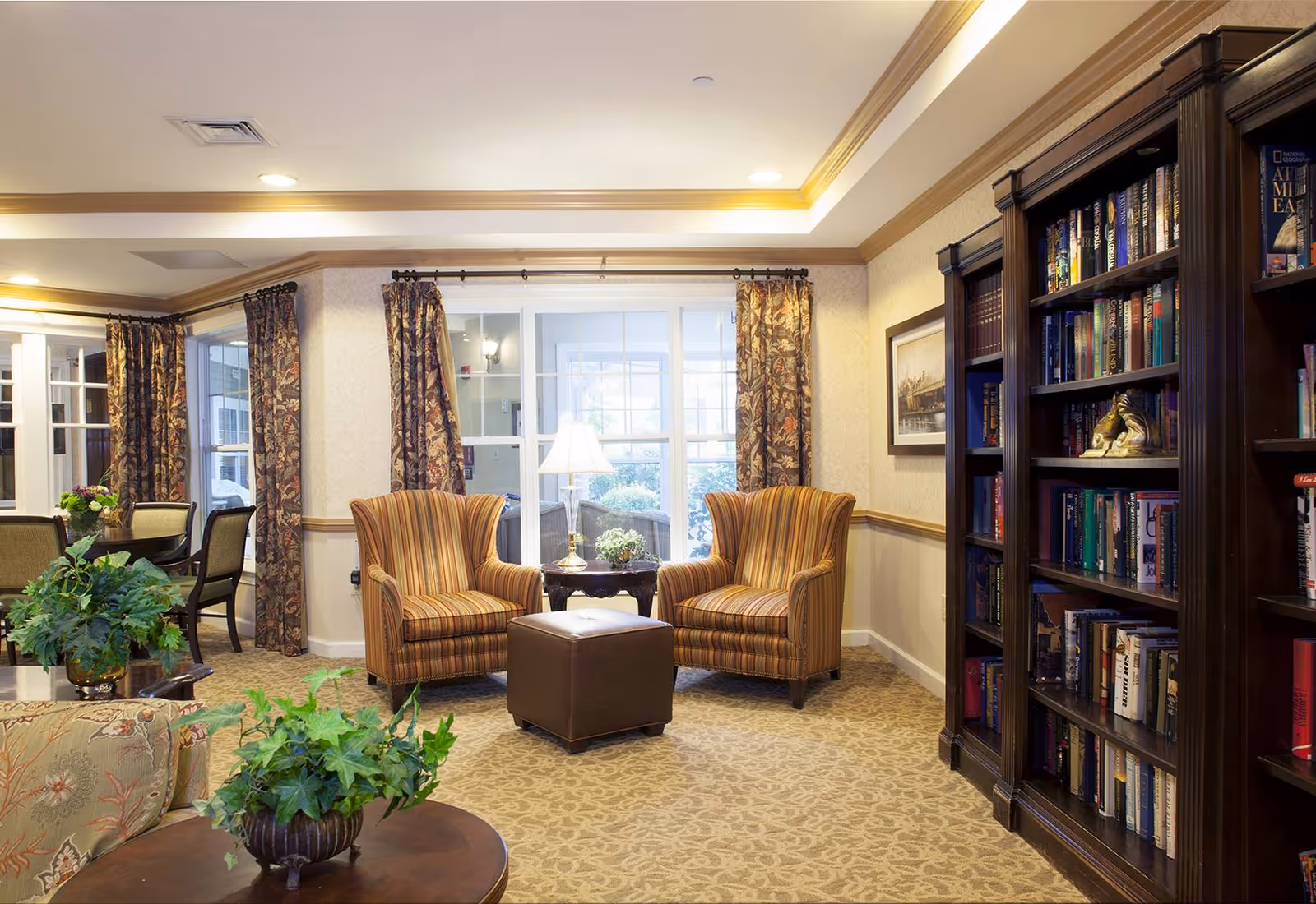 A cozy living room area in a senior living facility with two striped armchairs facing a brown ottoman, a wooden bookshelf filled with books, a round table with a green plant, and large windows with floral curtains letting in natural light.