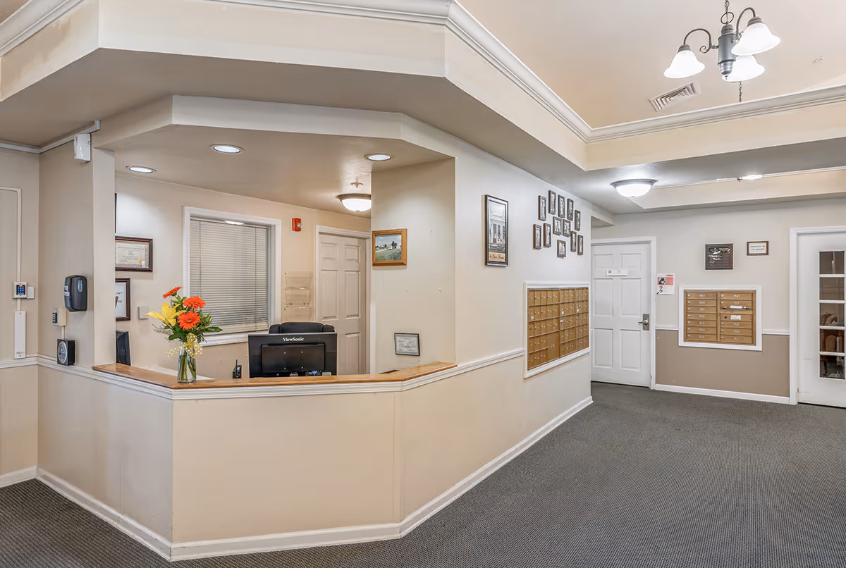 Reception desk and lobby area with a computer, vase of flowers, and wall mailboxes inside a senior living facility.