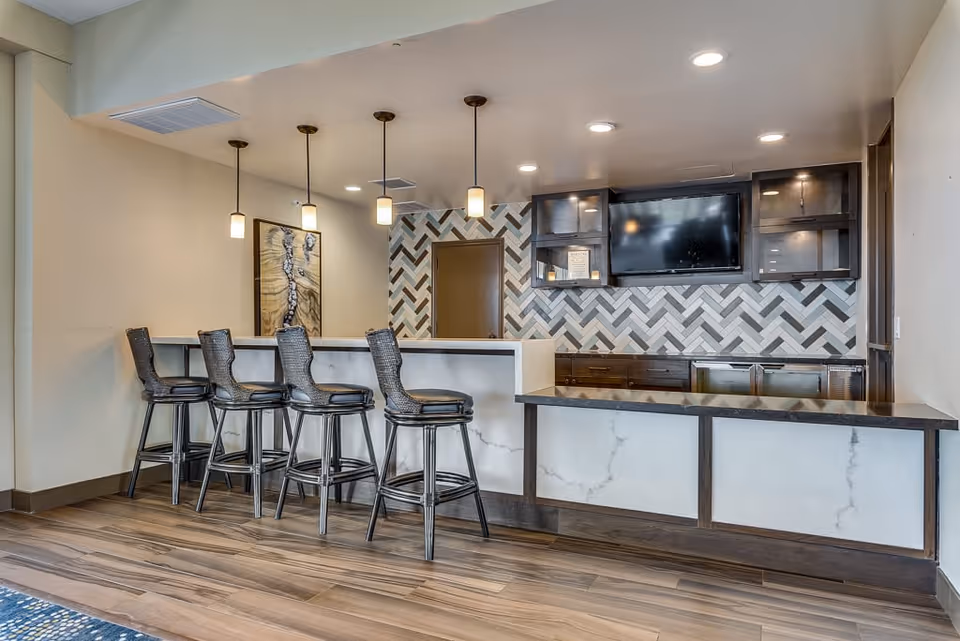 Modern kitchen area with a marble countertop bar and four high chairs. The back wall features a herringbone tile pattern with dark wood cabinets and a mounted flat-screen TV. Pendant lights hang from the ceiling above the bar, and the floor has wood-style tiles.