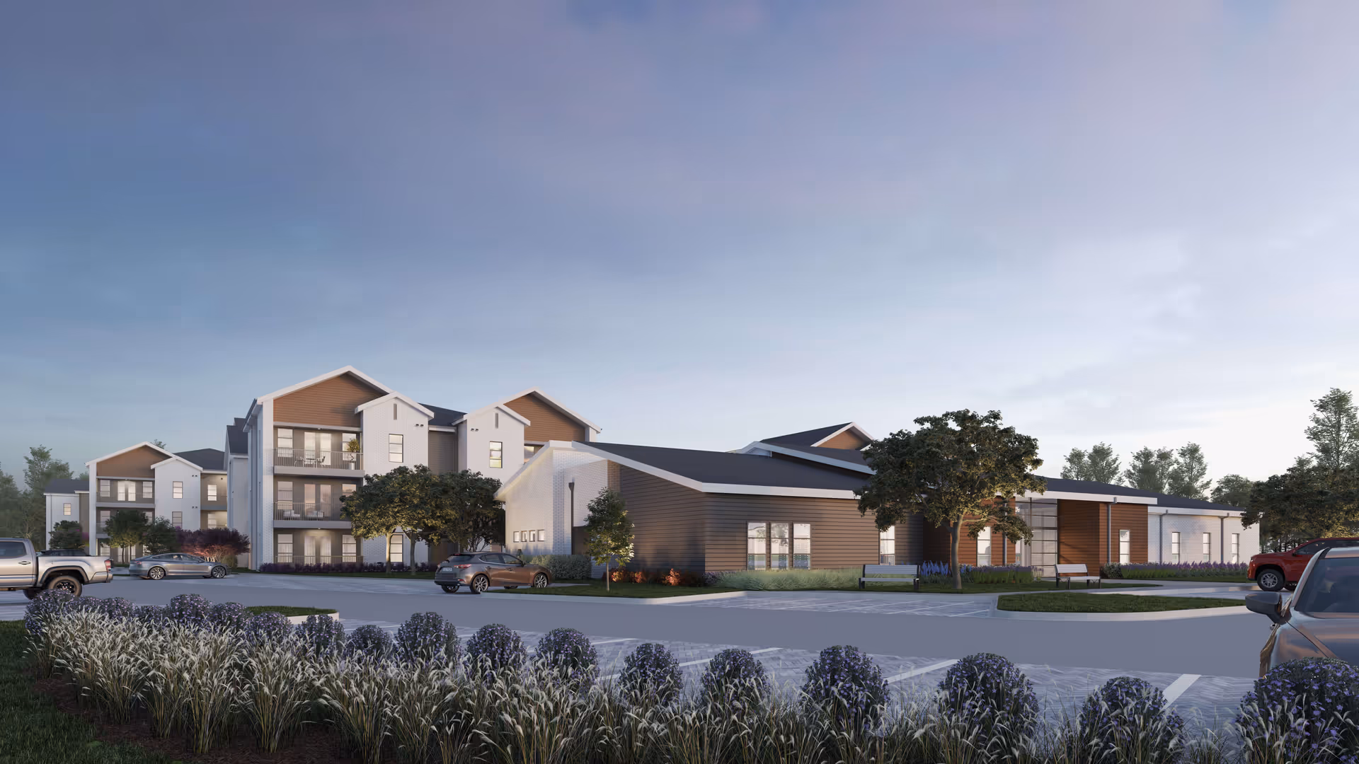 Exterior view of a modern senior living facility with multiple buildings, landscaped gardens, parked cars, and a clear sky at dusk.