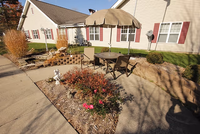 Outdoor patio area with a table, umbrella and chairs beside landscaped beds in front of a single-story building with red shutters.