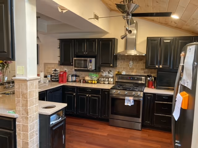 A modern kitchen with dark wooden cabinets, stainless steel appliances including a stove with a hood and a microwave, a refrigerator with papers attached, and a countertop with various kitchen items such as a coffee maker, spice rack, and jars. The floor is wooden and the ceiling has a wooden panel design.
