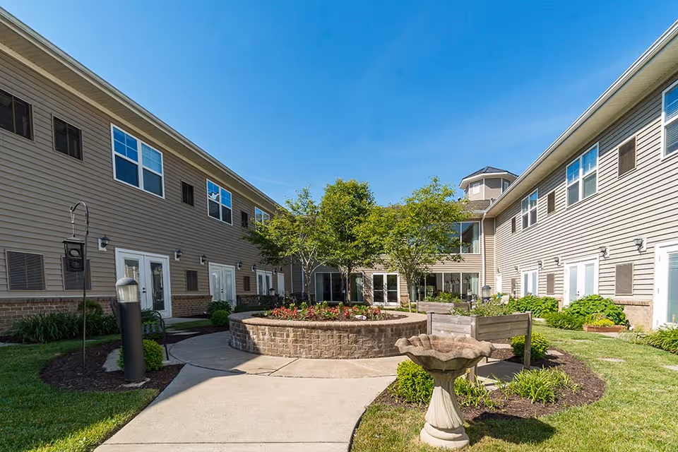 Outdoor courtyard area of a senior living facility with a circular raised flower bed in the center, surrounded by a paved walkway. The courtyard is flanked by two beige two-story buildings with multiple windows and doors. There are trees, green grass, and a birdbath in the foreground under a clear blue sky.