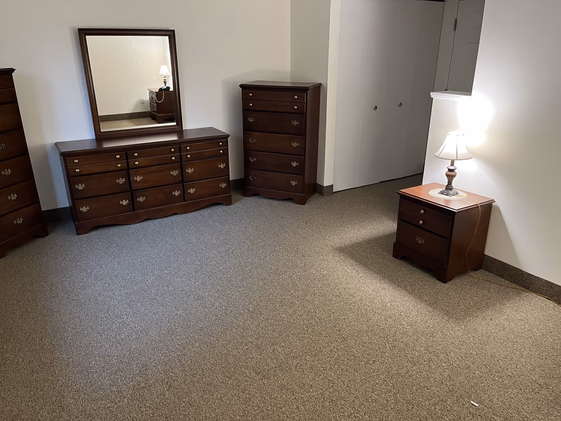 A room with beige carpet and white walls featuring wooden furniture including a dresser with a large mirror, a tall chest of drawers, and a nightstand with a lit table lamp. There is a closet with double doors in the corner.