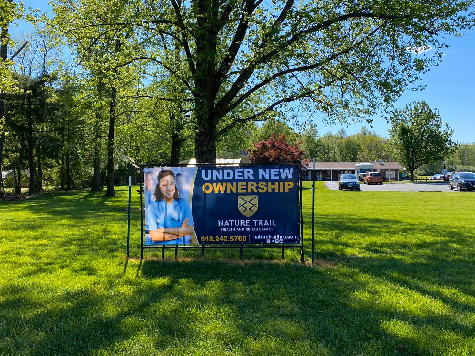 Green lawn and trees with a sign in front of a low building that reads "Under New Ownership" for Nature Trail Health and Rehab Center.