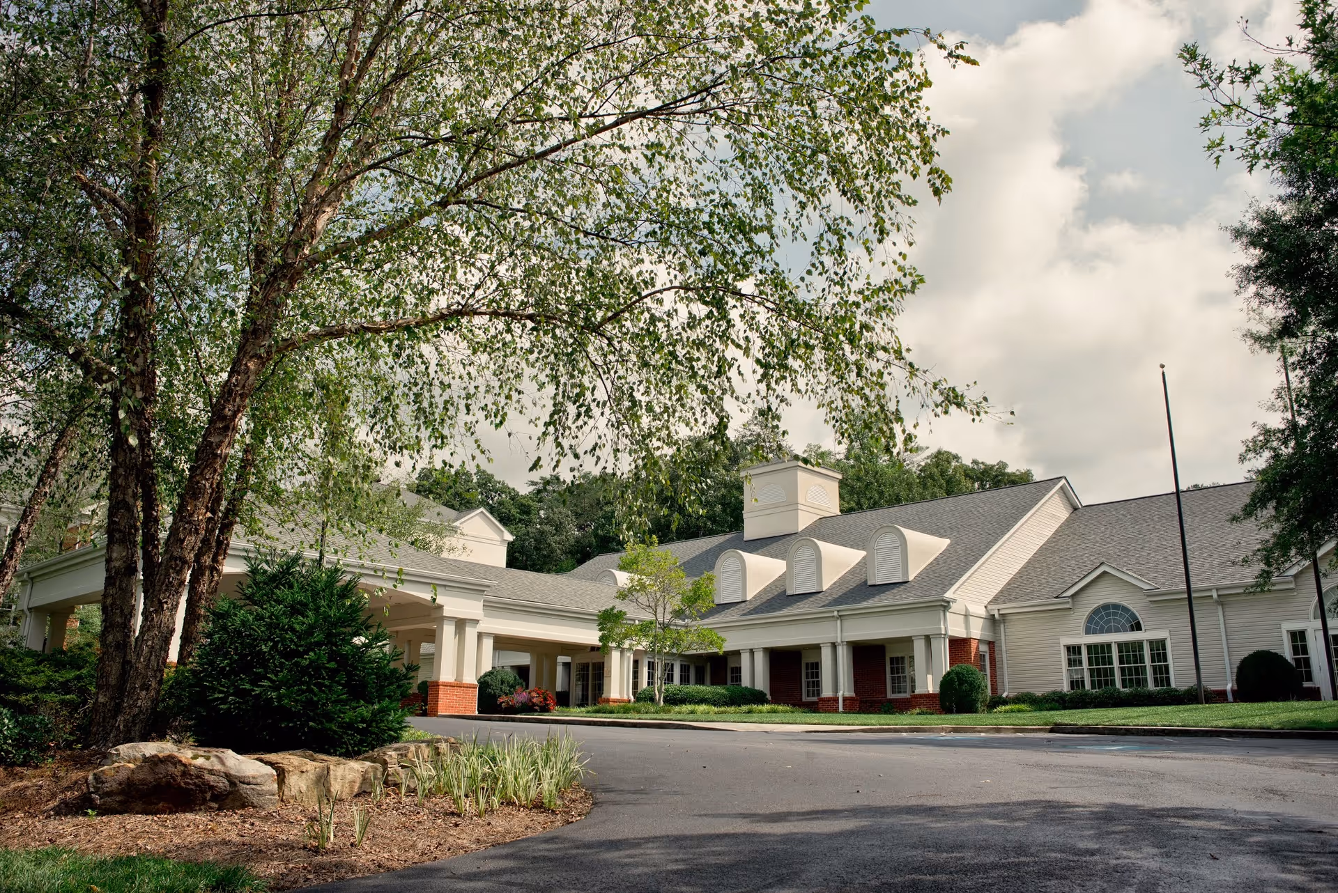 Exterior view of a single-story senior living facility building with white siding, red brick accents, and a covered entrance. The building is surrounded by trees, shrubs, and a paved driveway under a partly cloudy sky.