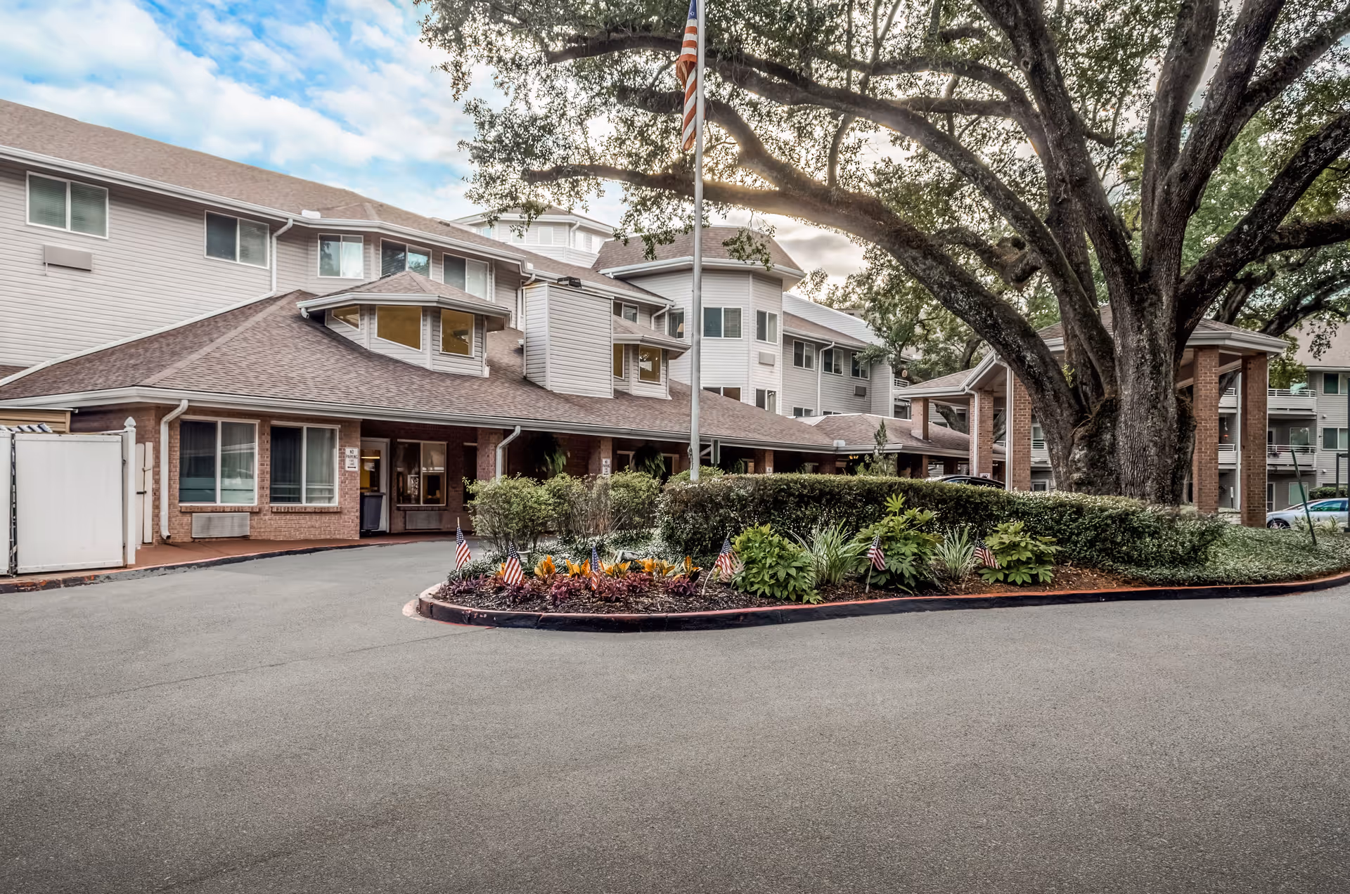 Front entrance and driveway of a multi-story senior living building with a landscaped circular planter and a large oak tree.