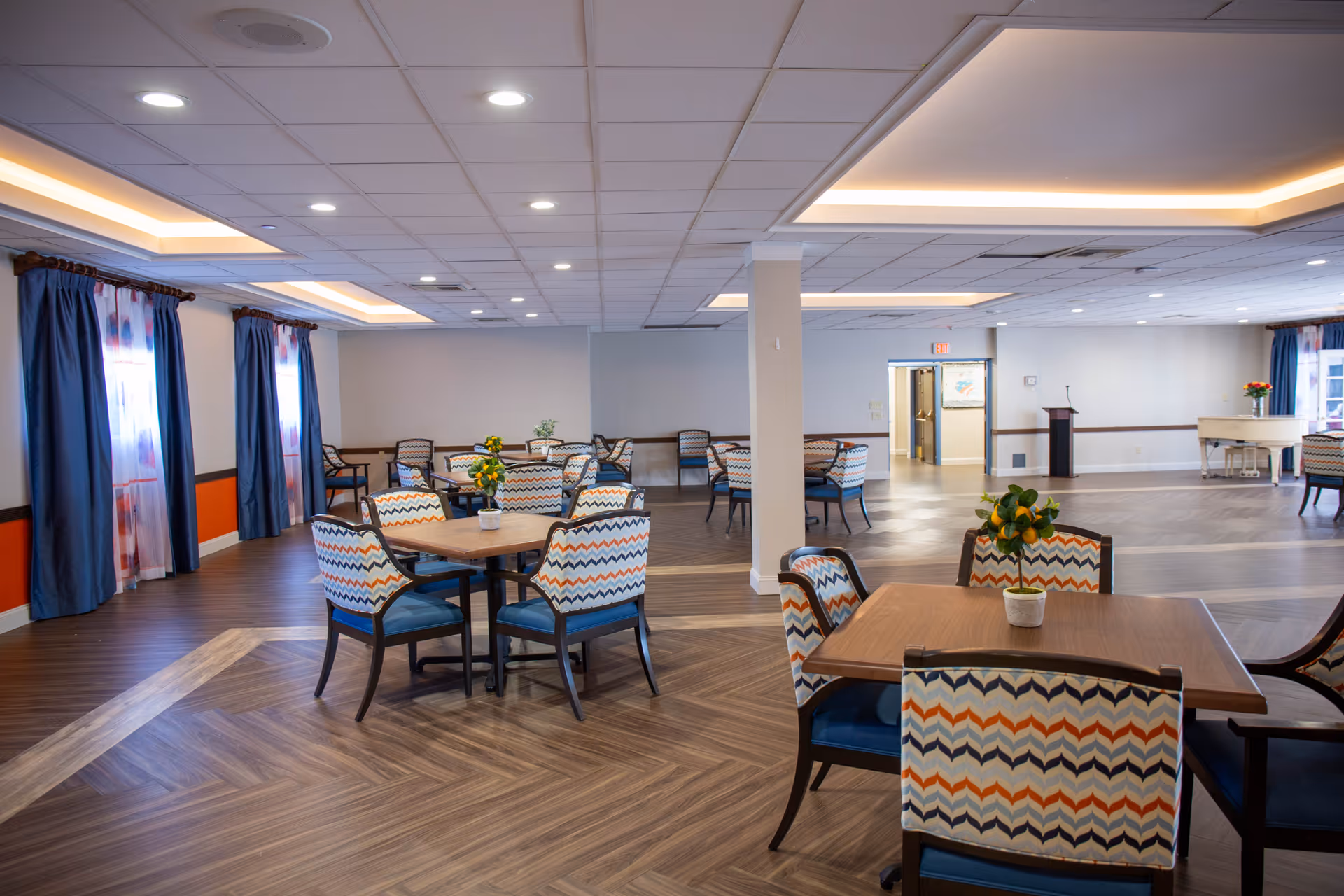 A spacious dining room with several wooden tables and chairs featuring colorful zigzag patterned upholstery. The room has large windows with blue curtains, wood-patterned flooring, and recessed ceiling lights. Small potted plants are placed on the tables, and a white piano is visible in the background near the far wall.