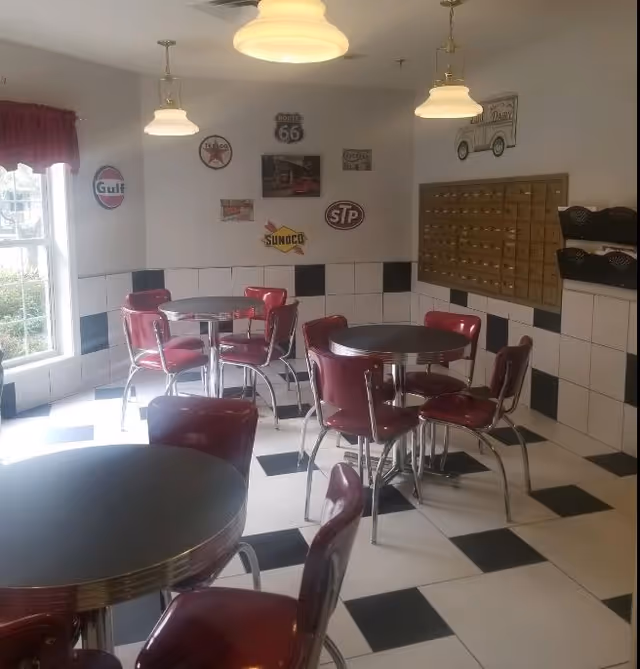 A retro-style dining area with black and white checkered floor and walls, featuring round tables with red cushioned chairs. The walls are decorated with vintage signs including Route 66, Texaco, Gulf, Sunoco, and STP. There are pendant lights hanging from the ceiling and a window letting in natural light.