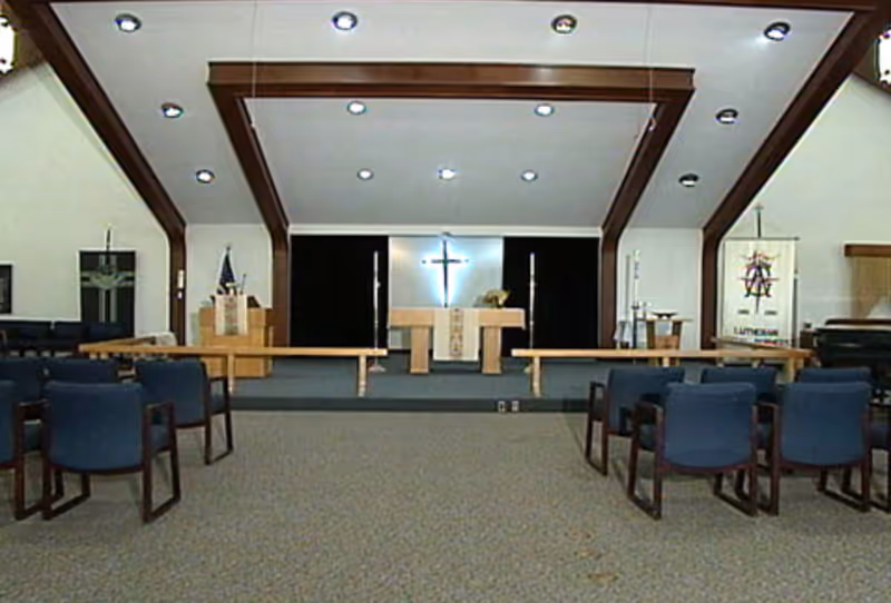 Interior chapel with an altar and illuminated cross at the front, wooden ceiling beams, and rows of blue chairs facing the platform.