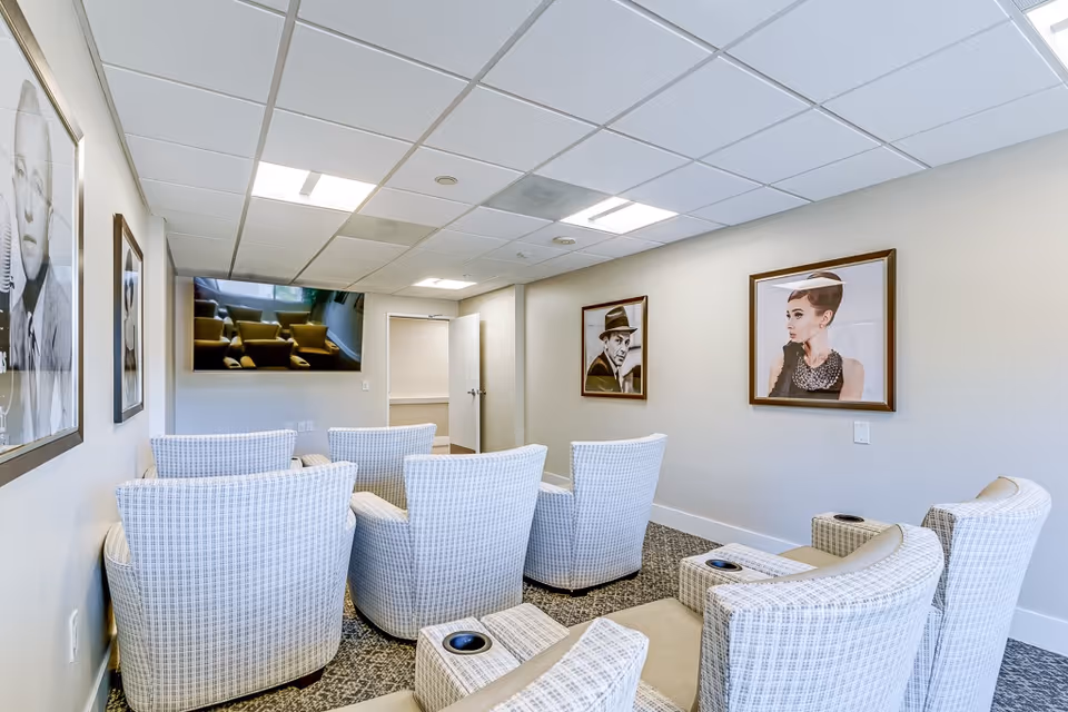 A small media room with eight cushioned armchairs arranged in two rows facing a wall-mounted flat-screen TV. The walls are decorated with framed black and white portraits of classic movie stars. The room has a drop ceiling with fluorescent lighting and a carpeted floor.