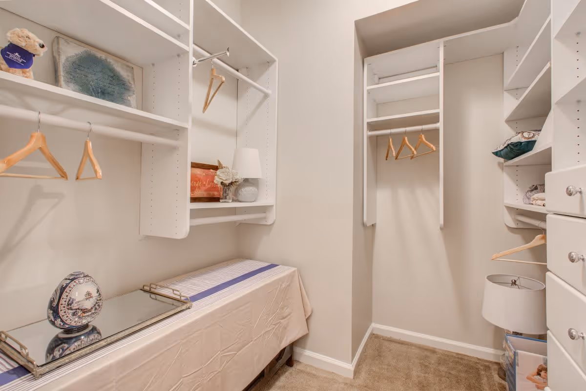 A walk-in closet with white built-in shelves and hanging rods. Several wooden hangers are hanging on the rods. The closet has a beige carpeted floor and light-colored walls. On the shelves and a covered surface below, there are decorative items including a lamp, framed pictures, and a decorative plate.