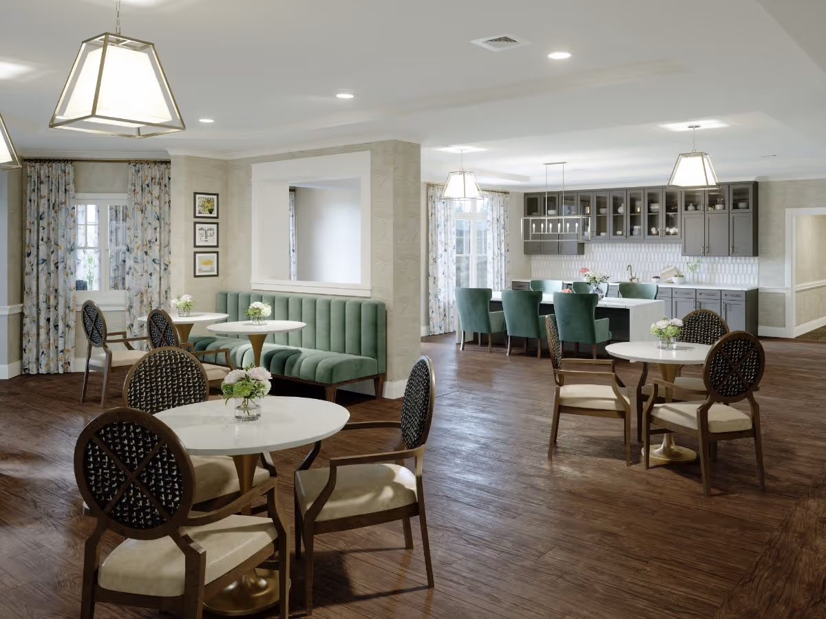 Bright communal dining area with round tables, upholstered chairs, green banquette seating, and a kitchen in the background.