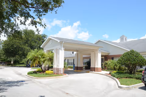 Covered porte-cochere entrance of a single-story senior living building with palm trees and landscaped grounds under a blue sky.