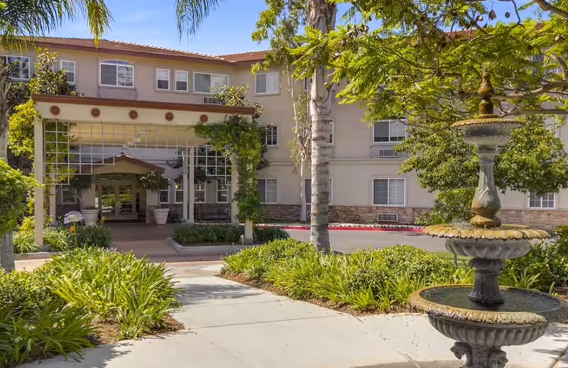Front entrance of a multi-story senior living building with a covered porte-cochere, landscaped walkway, and a tiered fountain.