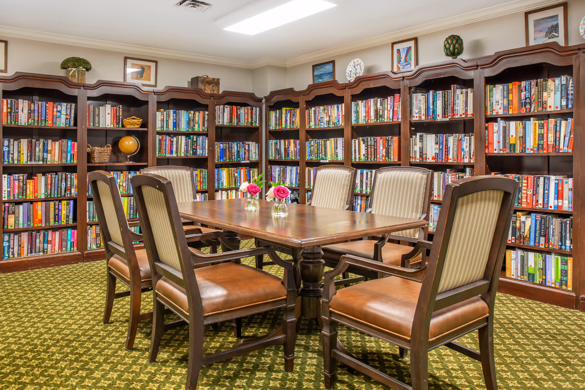 A cozy library room with wooden bookshelves filled with books lining the walls. In the center, there is a wooden table surrounded by six chairs with leather seats and striped fabric backs. Two small vases with pink and white flowers are placed on the table. The room has a green patterned carpet and framed artwork and decorative items on top of the bookshelves.