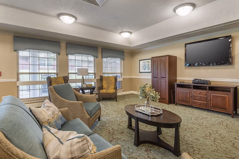 A cozy living room with a blue sofa, two armchairs, a wooden coffee table with a vase of flowers, and a wooden entertainment center with a flat-screen TV mounted on the wall. Three large windows with blinds and valances allow natural light into the room. The walls are beige with white trim, and the carpet has a green patterned design.
