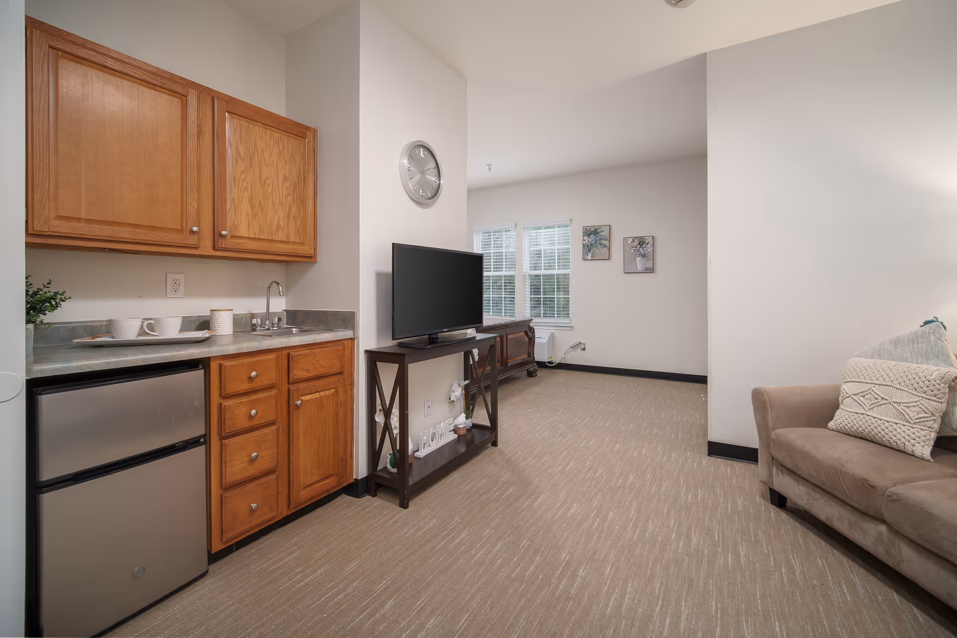 Interior view of a senior living facility room showing a small kitchenette with wooden cabinets, a mini refrigerator, and a sink on the left. In the center, there is a TV on a stand against a wall with a clock above it. On the right, a beige sofa with decorative pillows is partially visible. The room has beige carpet flooring and white walls with two framed pictures near a window in the background.