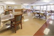 A spacious dining room in a senior living facility with multiple tables covered with tablecloths and surrounded by wooden chairs. The room has large windows allowing natural light to fill the space, and the floor is a polished linoleum with a reddish border. The walls are light-colored with some decorations and a bulletin board visible.