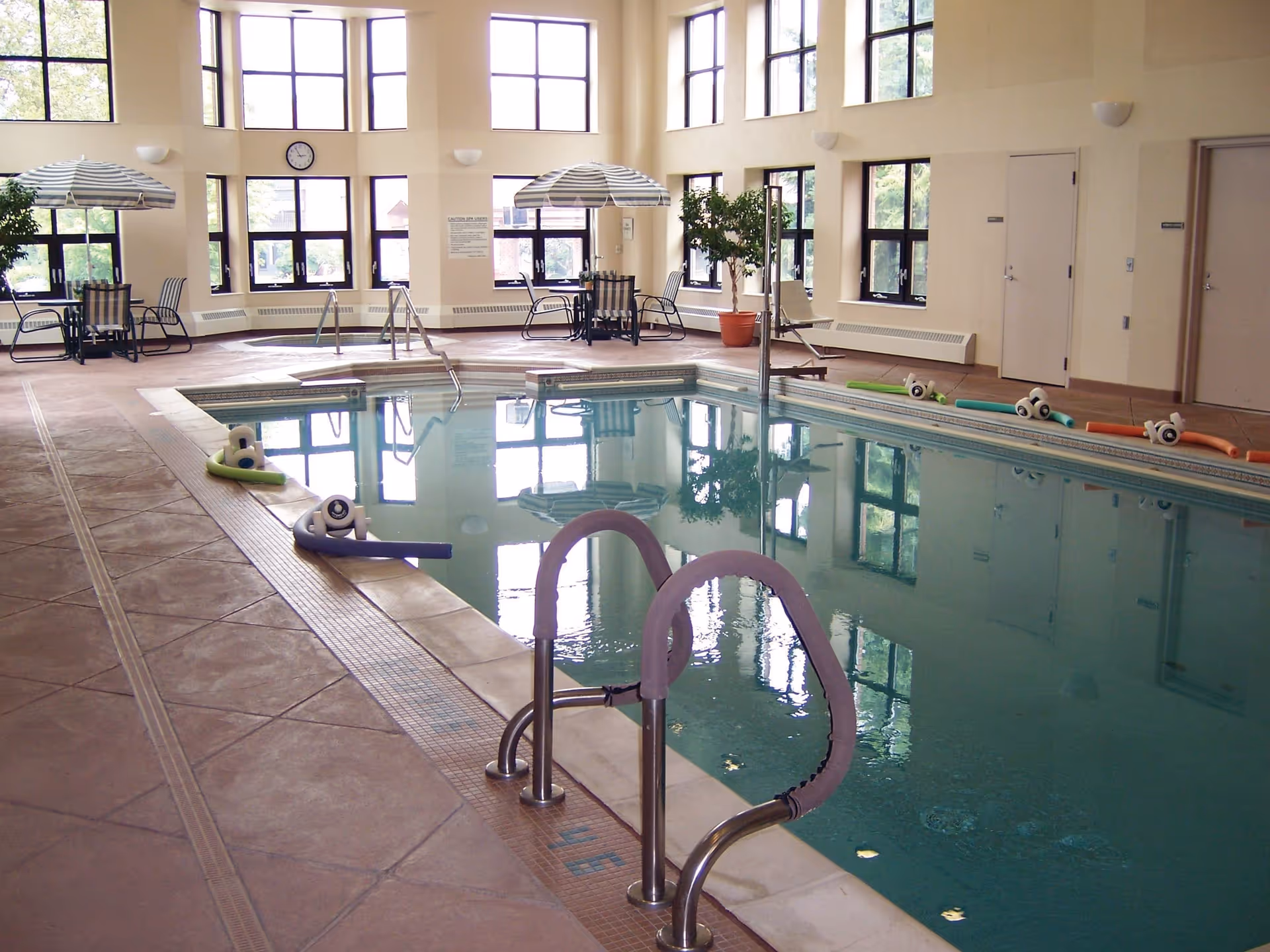 Indoor swimming pool area with large windows letting in natural light, poolside chairs with umbrellas, pool noodles and flotation devices around the pool, and a clock on the wall.