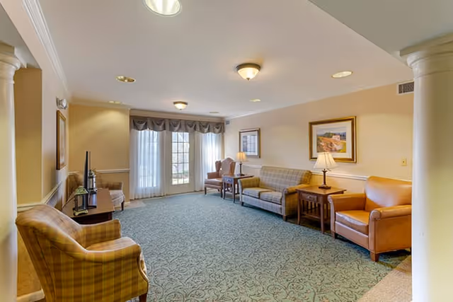 Spacious senior living common room with sofas, armchairs, side tables, lamps and large windows with curtains.