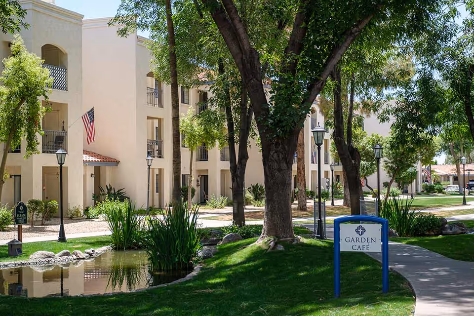 Outdoor view of Fellowship Square Phoenix senior living facility showing a landscaped garden area with trees, a small pond, and a pathway. Beige multi-story buildings with balconies and American flags are visible in the background. A sign reading 'Garden Café' is placed near the pathway.