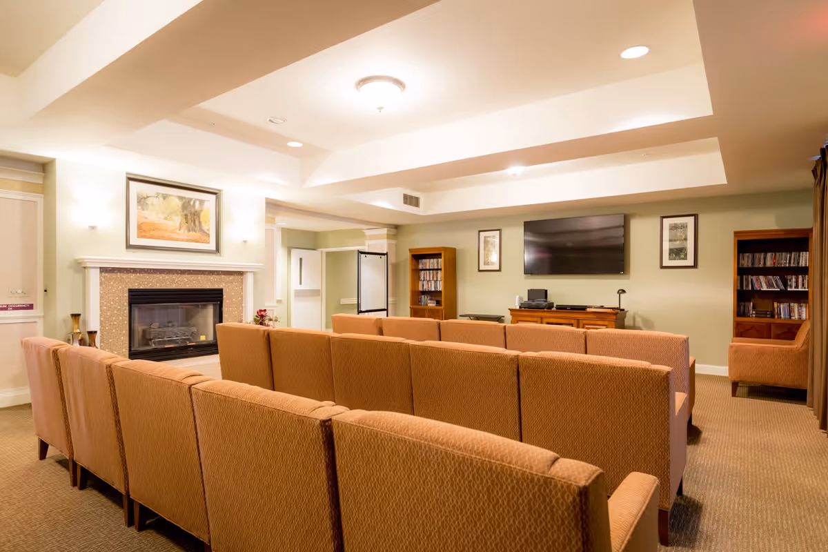 A cozy common area with rows of orange upholstered chairs facing a wall-mounted flat-screen TV. The room features a fireplace with a framed painting above it, bookshelves filled with DVDs or books, and soft lighting from ceiling fixtures.