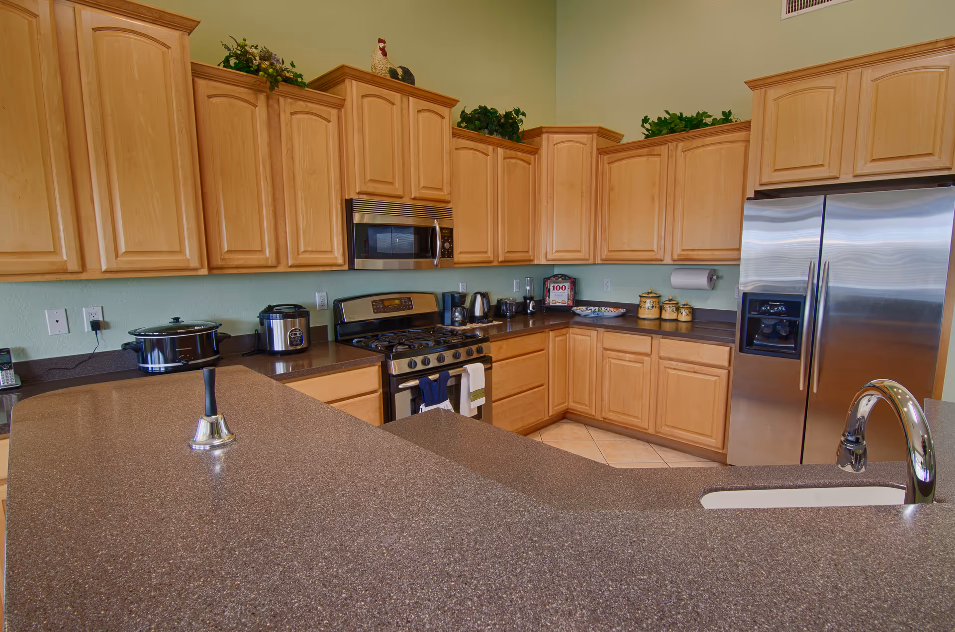 A spacious kitchen with light wood cabinets, a stainless steel refrigerator, a gas stove with an overhead microwave, and various small kitchen appliances on the countertops. There is a large brown countertop with a sink and a shiny faucet in the foreground, and decorative greenery on top of the cabinets.